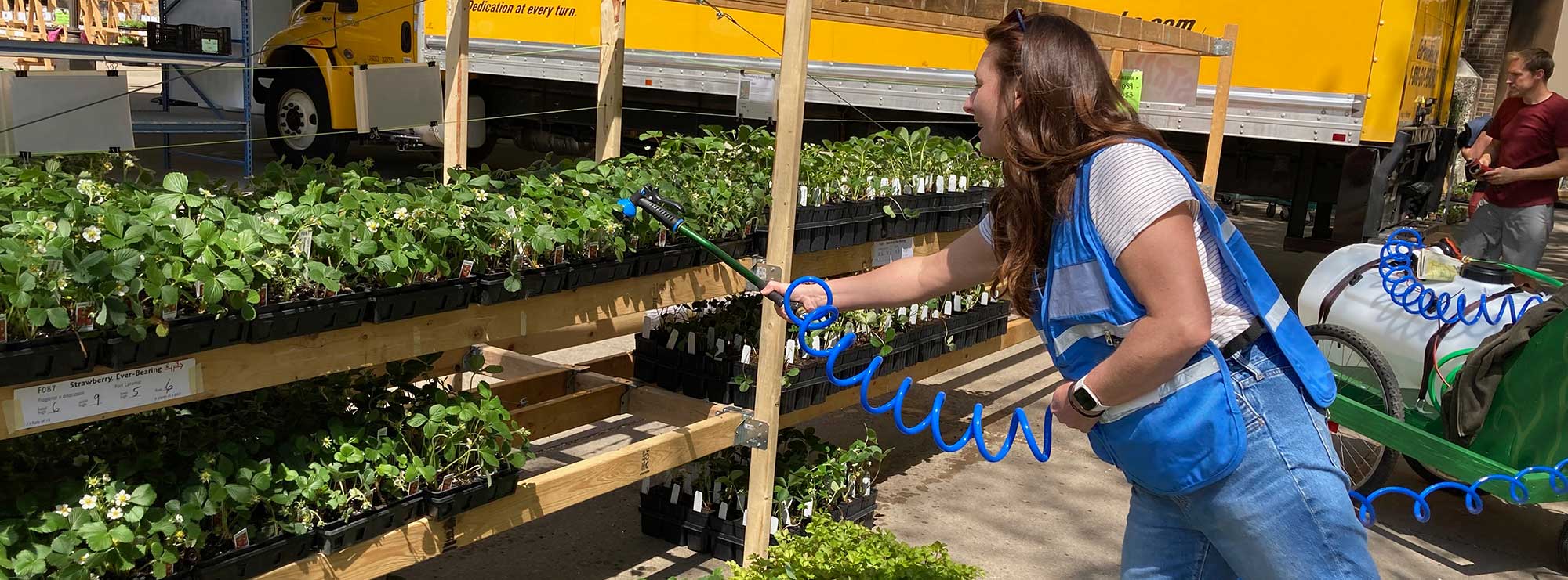 A woman in a blue safety vest waters a table full of plants with a gardening wand attached to a large watering cart. Yellow rental truck in background.